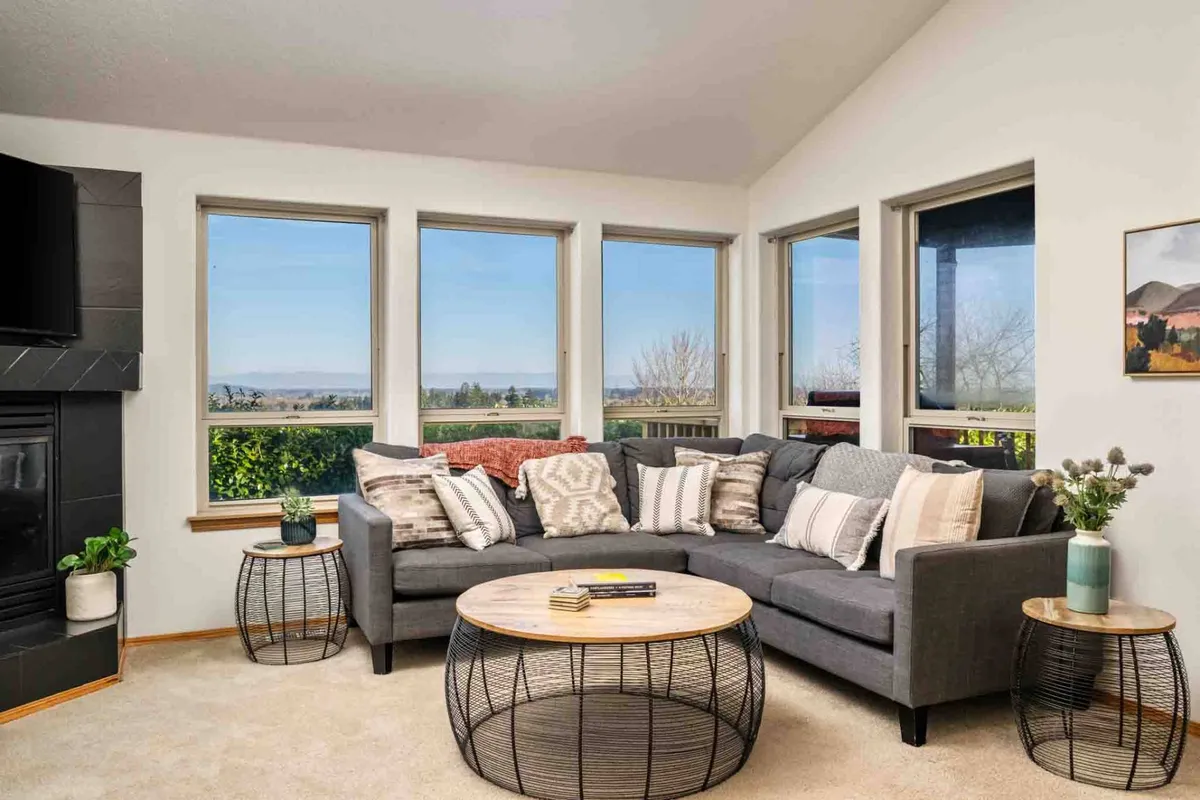 A large sectional sofa, coffee table, and windows with scenic views create a cozy, welcoming atmosphere ideal for lounging. The room is filled with natural light and offers ample space for both people and pets to gather and relax.