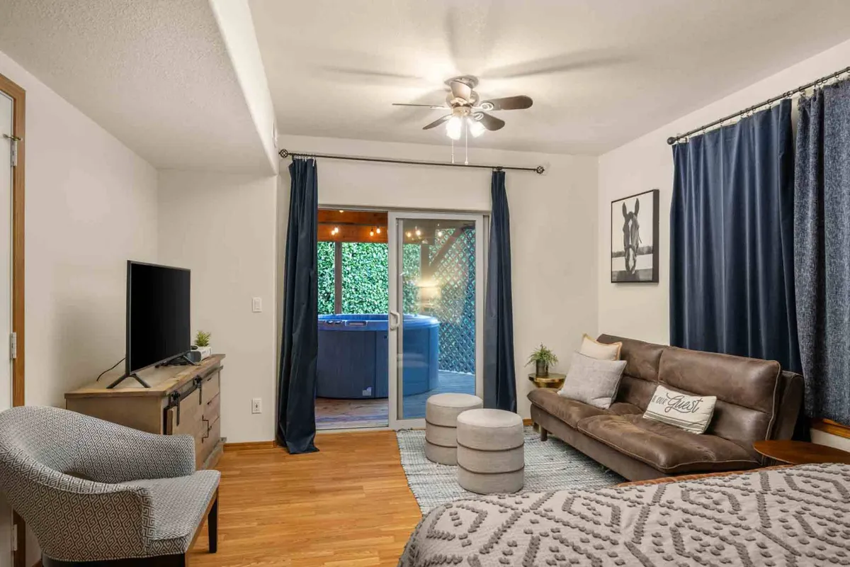 This image shows a cozy living room with a plush sofa, television, decorative pillows, and a sliding glass door leading to an outdoor area. Through the door, a hot tub is visible, suggesting a seamless connection between indoor comfort and backyard relaxation.