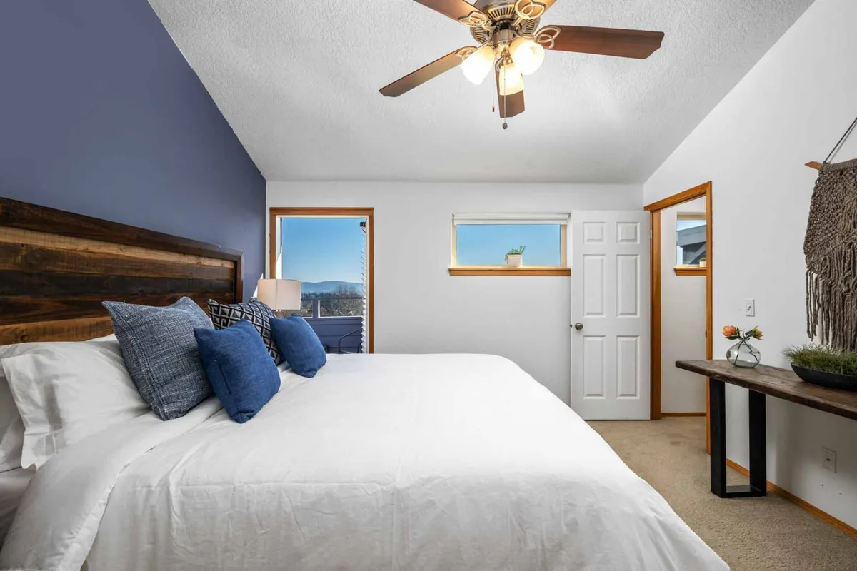A serene bedroom featuring a wooden headboard, blue accent wall, and crisp white linens. There's a ceiling fan for comfort and a window letting in natural light, making the space inviting for rest and relaxation.