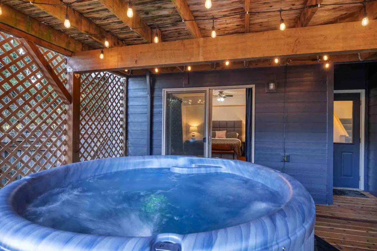 An inviting hot tub sits on a deck, framed by string lights and an open bedroom door, with views stretching out over the valley. The setting offers relaxation and a seamless transition between indoor comfort and outdoor splendor.