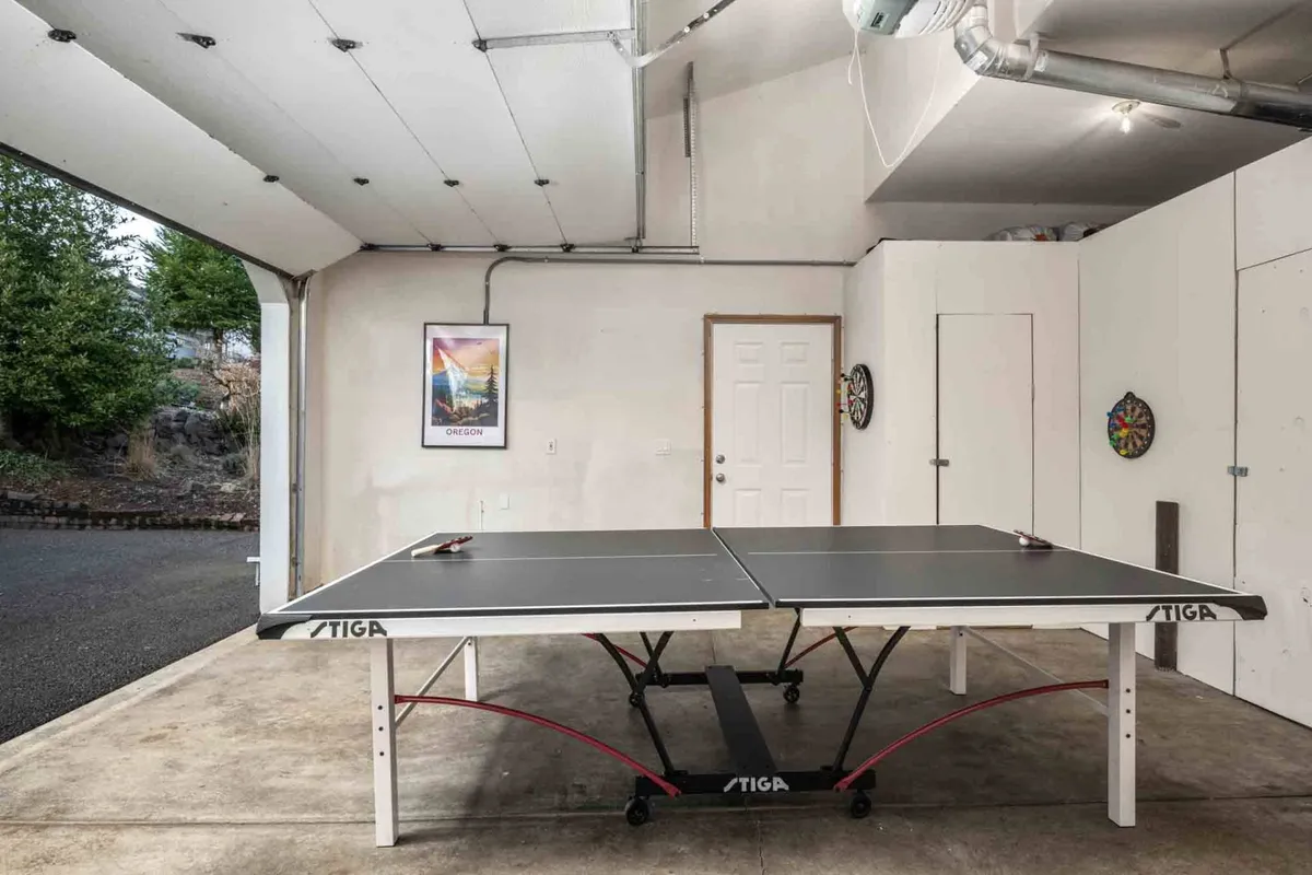 This image showcases the garage game room with a ping pong table, dartboard, and door leading to the outside. The walls are decorated with artwork, and the space is bright and inviting, clearly designed for group games and socializing.