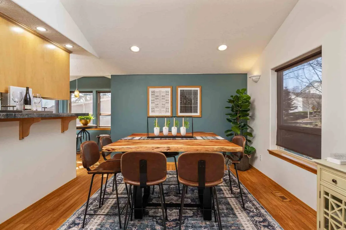 A cozy dining area with a wooden table, striking green accent wall, lush plant, and eye-catching lighting fixtures. Windows provide glimpses of the outdoors, and the setup suggests a perfect spot for group meals or festive gatherings.
