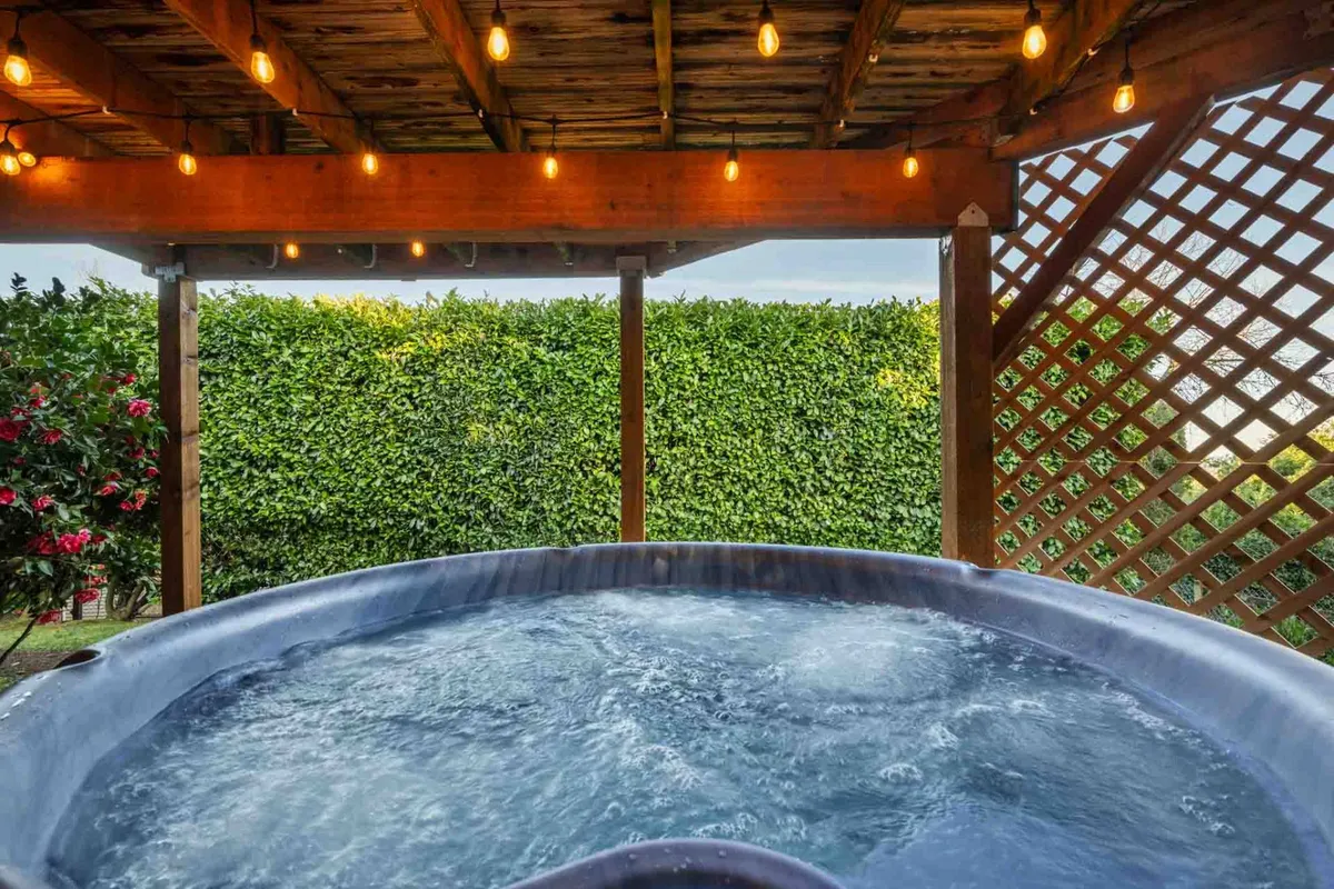 The image features a hot tub set under a wooden pergola with string lights, surrounded by lush greenery. The inviting ambiance is perfect for evening relaxation and social gatherings, aligning with the description of the hot tub under bistro lights on the lower deck.