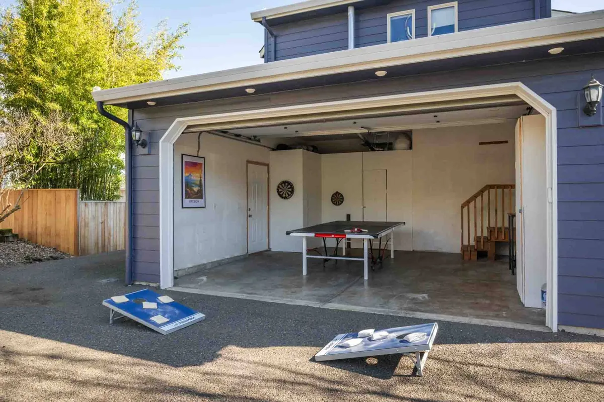 The image displays a ping pong table and dartboard set up in a covered exterior or partially open space, with a staircase and plenty of room for group games. The area feels energetic and ready for active fun, possibly accommodating cornhole as well.