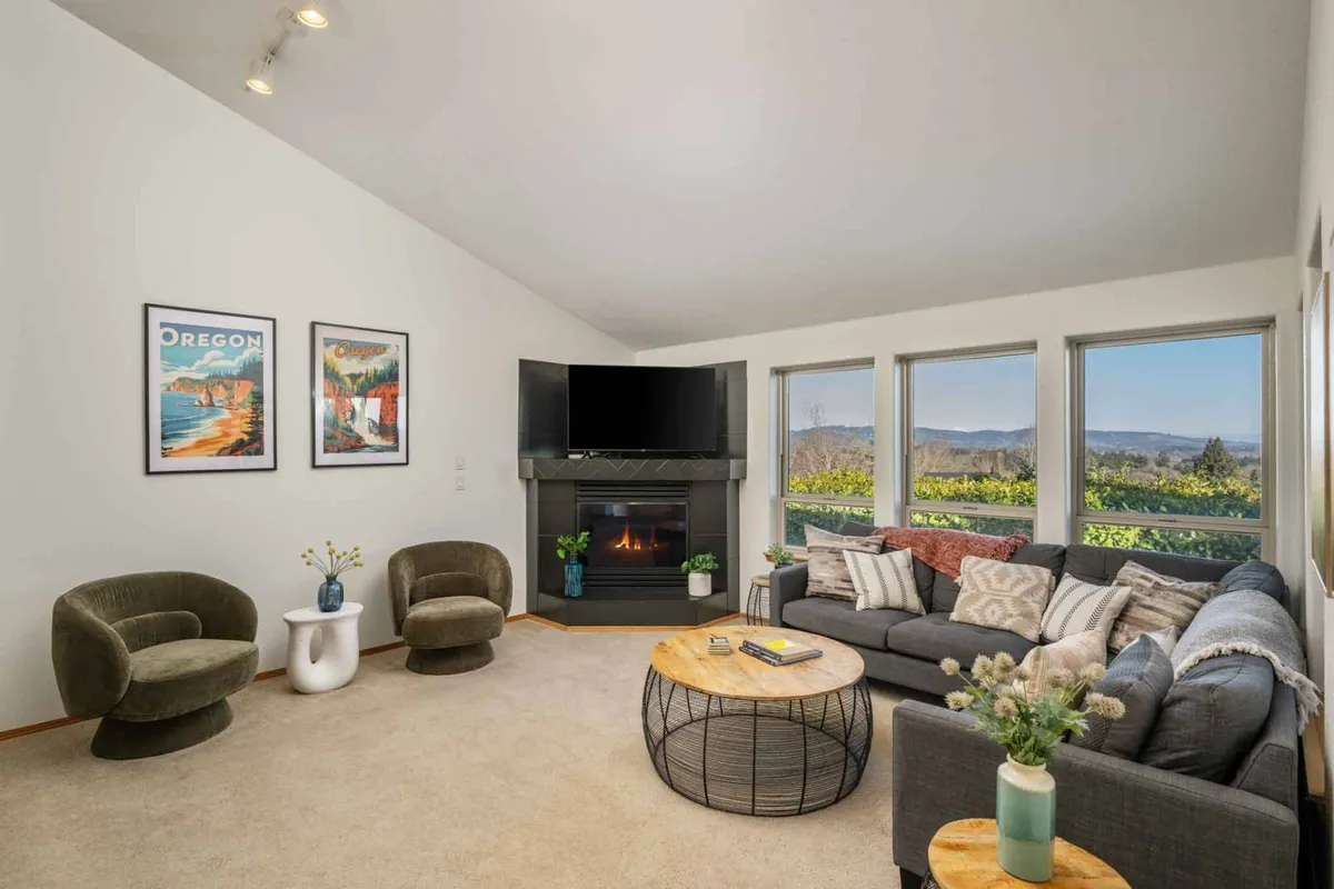 The image showcases a welcoming living room with a modern fireplace as the centerpiece, surrounded by a comfortable sofa, stylish coffee table, and tasteful artwork. Expansive windows frame breathtaking sunset views over the Willamette Valley, filling the room with a warm glow and inviting natural light. The setting is perfect for relaxing, socializing, or simply soaking in the serene Oregon wine country atmosphere.